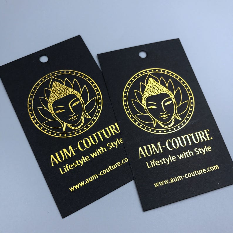 Pu&ograve; includere: Due etichette regalo nere con lettering dorato e un'immagine dorata di una testa di Buddha. Il testo sulle etichette recita "AUM-COUTURE Lifestyle with Style" e "www.aum-couture.co".