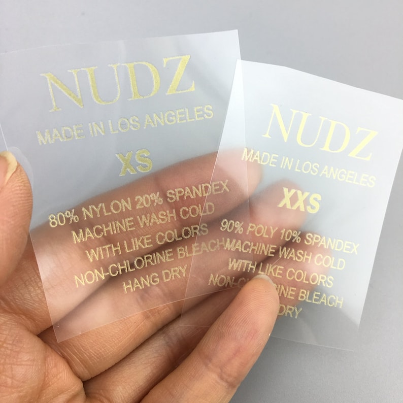 May include: Two clear plastic clothing labels with gold lettering. The labels are for "NUDZ" clothing and state "Made in Los Angeles". One label is for size "XS" and the other is for size "XXS". Both labels include care instructions.