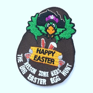 May include: A brown embroidered patch featuring a green bird wearing a pirate hat and holding a sign that reads "Happy Easter". The text surrounding the bird says "The Nelson Zone Has Big Easter Egg Hunt".