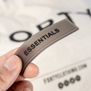 May include: A gray fabric label with the word "ESSENTIALS" printed in black. The label is curved and has a slight sheen.