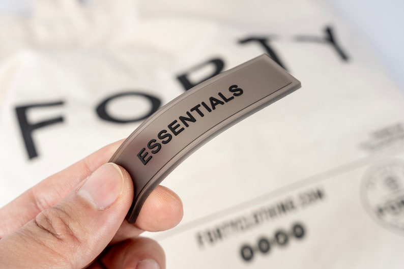 May include: A gray fabric label with the word "ESSENTIALS" printed in black. The label is curved and has a slight sheen.