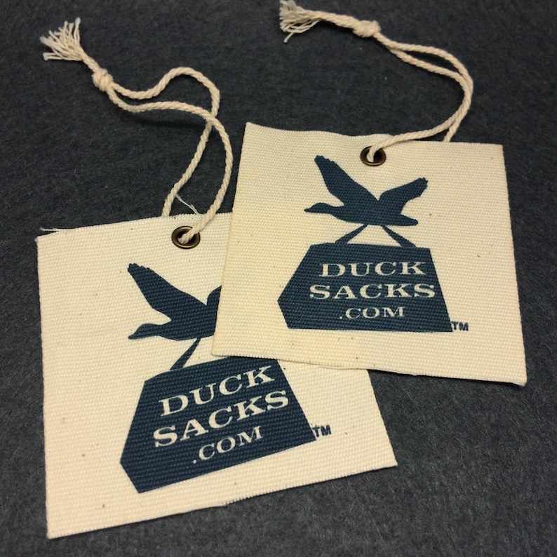 Puede incluir: Dos etiquetas de tela blanca con un logotipo de pato azul y el texto "Duck Sacks .com" y un s&iacute;mbolo de marca registrada. Las etiquetas est&aacute;n unidas a una cuerda.