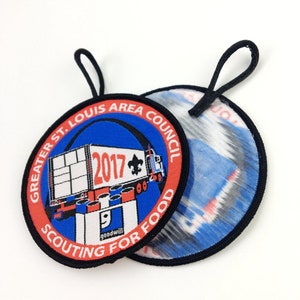 May include: A red, white, and blue embroidered patch with the text "GREATER ST. LOUIS AREA COUNCIL" and "SCOUTING FOR FOOD" around a blue and white illustration of a truck with the text "2017" on the side. The patch has a black border and a black loop on the top.