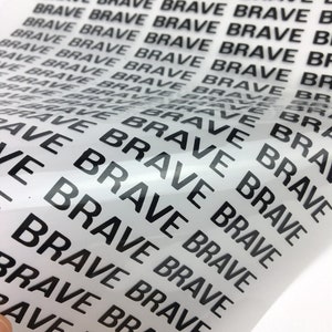 以下が含まれることがあります： 透明なビニールシートのクローズアップ。黒いインクで「BRAVE」という単語が繰り返し印刷されています。