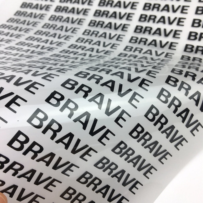 以下が含まれることがあります： 透明なビニールシートのクローズアップ。黒いインクで「BRAVE」という単語が繰り返し印刷されています。