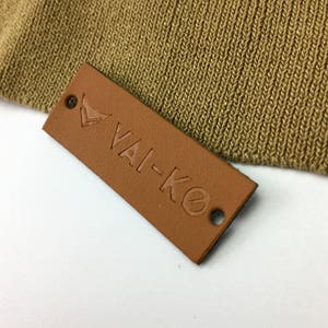 May include: A brown leather label with the text "VAI-KO" embossed in a darker brown color. The label has two small holes on the top and bottom edges.