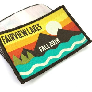 May include: A brown, rectangular patch with a colorful sunset design. The patch features a white sun, brown mountains, green trees, and a blue lake with white waves. The text "FAIRVIEW LAKES FALL 2018" is printed on the patch.