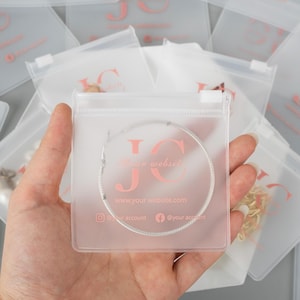 May include: Clear plastic jewelry bags with a pink logo and website information. A silver bracelet is visible inside one bag. Other bags with jewelry are in the background.