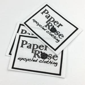 May include: Close-up of three white fabric labels with black text and a black rose graphic. The text reads "Paper Rose" and "Upcycled Clothing".