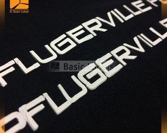 May include: Close-up of a black fabric with white raised lettering that reads "FLUGERVILLE" twice. The lettering is slightly distressed.