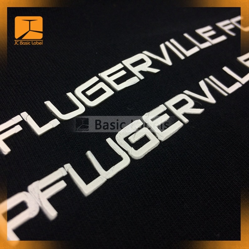 May include: Close-up of a black fabric with white raised lettering that reads "FLUGERVILLE" twice. The lettering is slightly distressed.