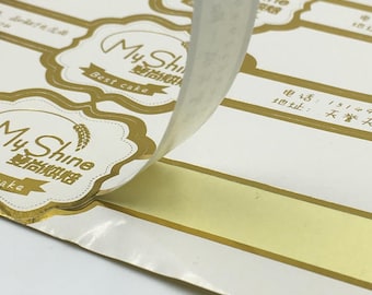 Może przedstawiać: Naklejki z folii złotej z tekstem "My Shine" i "雙尚烘焙" w chińskich znakach. Naklejki są przeznaczone do produktów piekarniczych i mają złotą ramkę oraz wzór pszenicy. Tekst "Best cake" jest również umieszczony na naklejce.