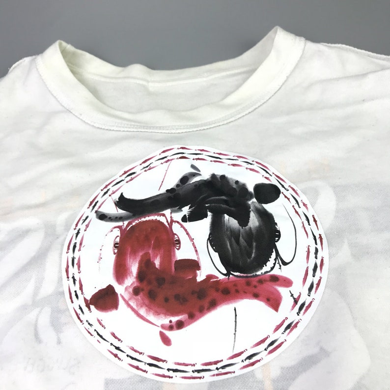 May include: White t-shirt with a circular design featuring two fish, one black and one red, painted in a watercolor style.