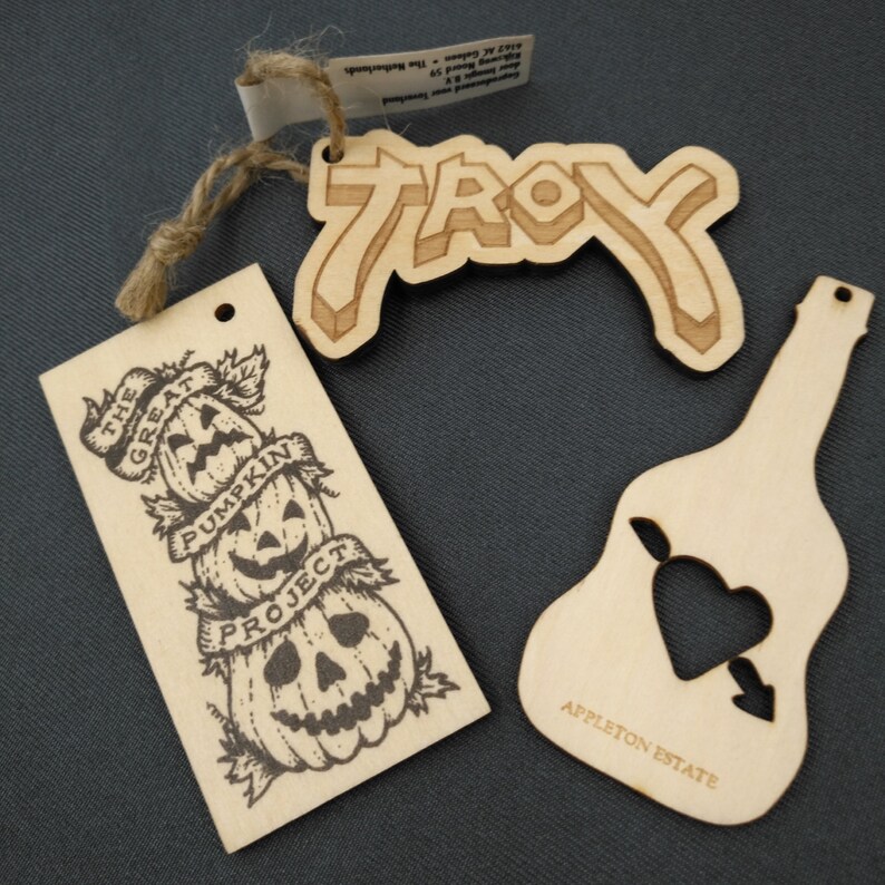 May include: Three wooden ornaments with laser-cut designs. The first ornament is a rectangular tag with the text "The Great Pumpkin Project" and a stack of three jack-o'-lantern faces. The second ornament is a curved tag with the text "TROY" in a stylized font. The third ornament is shaped like a guitar with a heart and arrow cut out and the text "APPLETON ESTATE".