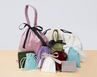 May include: A collection of velvet and satin drawstring bags in various colors, including pink, green, purple, and white. Each bag is tied with a contrasting ribbon, and some have handles. A small, rectangular gray tag is also present.