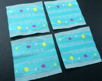 May include: Four turquoise blue fabric labels with white stitching. Each label has a white design of a string of lights with yellow and purple circles. The text "RubyLOLA design" is written in white on each label.