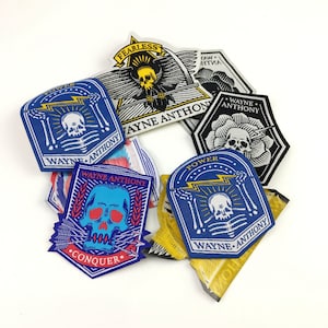 Puede incluir: Una colección de nueve parches bordados con un motivo de calavera. Los parches son azules, negros, blancos, amarillos y rojos. Los parches tienen el texto "WAYNE ANTHONY", "FEARLESS", "POWER" y "CONQUER" en ellos.