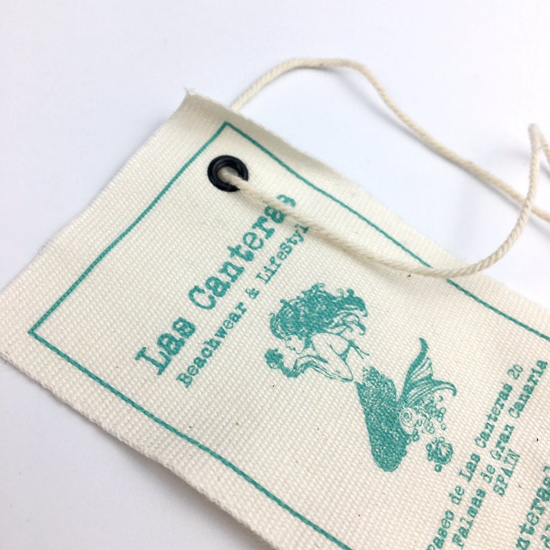 Puede incluir: Una etiqueta de lona blanca con un borde turquesa y una ilustraci&oacute;n de sirena turquesa. La etiqueta tiene un ojal negro y una cuerda atada. El texto en la etiqueta dice "Las Cantera Beachwear & Lifestyle" y "Paseo de Las Canteras 20 Palmas de Gran Canaria ESPA&Ntilde;A".