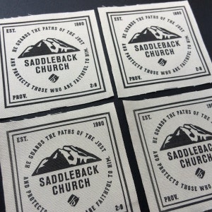 May include: Four beige fabric patches with black text and a mountain graphic. The text reads "EST. 1980 HE GUARDS THE PATHS OF THE JUST AND PROTECTS THOSE WHO ARE FAITHFUL TO HIM. SADDLEBACK CHURCH PROV. 2:8".
