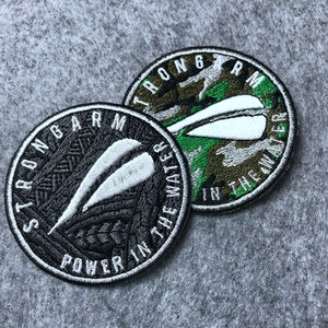 May include: Two embroidered patches with a camouflage background and white text. The text on the top patch reads "Strongarm in the water". The text on the bottom patch reads "Strongarm Power in the water".
