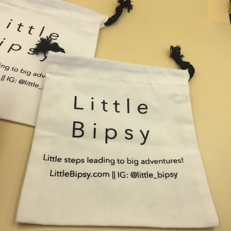 May include: Two white cotton drawstring bags with black text. The top bag says "little ipsy" and the bottom bag says "Little Bipsy" and "Little steps leading to big adventures! LittleBipsy.com || IG: @little_bipsy".