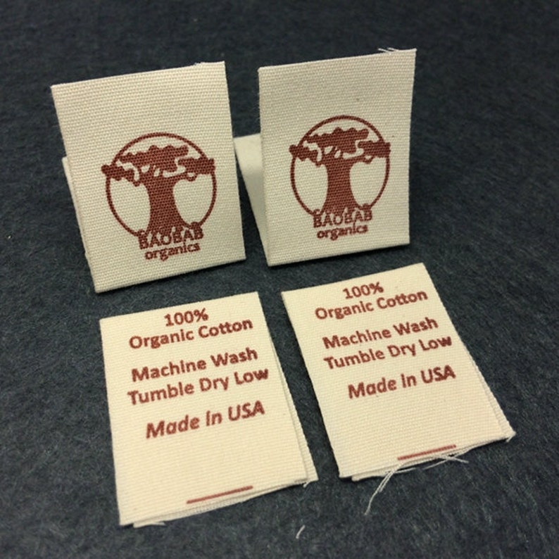 May include: Four white fabric labels with brown text and a brown tree logo. The labels read "100% Organic Cotton", "Machine Wash", "Tumble Dry Low", and "Made in USA". The logo is a circle with a tree inside and the words "BAOBAB organics" underneath.