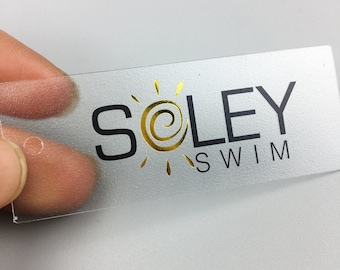 May include: A clear plastic tag with a gold sun and the text "SOLEY SWIM" in black.