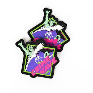 May include: Two black, square stickers with a graphic design. The design features the Statue of Liberty riding a wave, with the words "CRIME WAVE" in pink and the website address "OFFDUTYSTICKERS.COM". The background is purple and green.