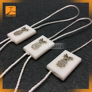 May include: Three white plastic string tags with gold pineapple designs. The tags are attached to white string.