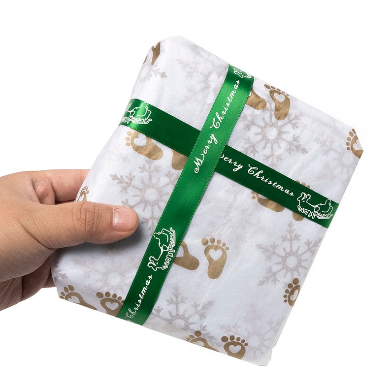 以下が含まれることがあります： 緑のリボンが結ばれた白いギフトボックス。リボンには「メリークリスマス」の文字が印刷されています。ボックスは雪片と足跡のパターンで飾られています。