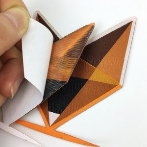 May include: A geometric iron-on patch with a brown, orange, and black design. The patch has a white backing that can be peeled off before ironing.