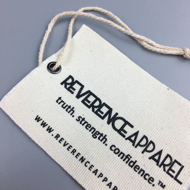 Puede incluir: Una etiqueta de tela de algod&oacute;n blanco con texto negro que dice "REVERENCE APPAREL truth. strength. confidence. TM" y "WWW.REVERENCEAPPAREL.COM". La etiqueta tiene un ojal de metal y una cuerda blanca atada.