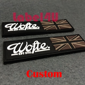 Könnte beinhalten: Zwei schwarze und braune Gummipatchen mit dem Text "Wofte. EST 2011." und einem Union-Jack-Design. Die Patches befinden sich auf einer braunen Oberfläche.