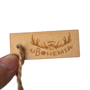 May include: A wooden tag with a brown, natural finish. The tag has a hole for string and features a laser-engraved design of antlers, an eye, and the text "UBOHEMIA" with the year "MMXVI" above the eye.