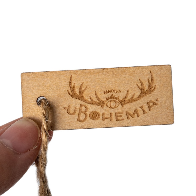 May include: A wooden tag with a brown, natural finish. The tag has a hole for string and features a laser-engraved design of antlers, an eye, and the text "UBOHEMIA" with the year "MMXVI" above the eye.