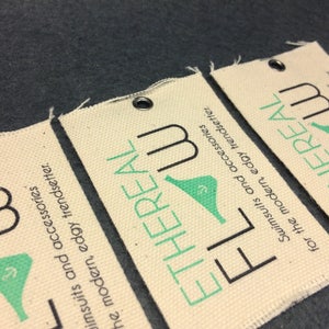 Puede incluir: Dos etiquetas de tela blanca con texto negro y verde. El texto en las etiquetas dice "ETHEREAL" y "FLOW". Las etiquetas son para trajes de ba&ntilde;o y accesorios.