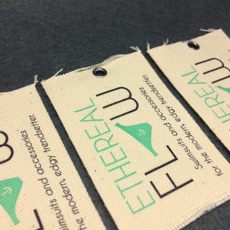 Puede incluir: Dos etiquetas de tela blanca con texto negro y verde. El texto en las etiquetas dice "ETHEREAL" y "FLOW". Las etiquetas son para trajes de ba&ntilde;o y accesorios.