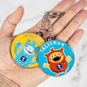 Puede incluir: Dos llaveros redondos con esmalte azul y amarillo. Cada llavero presenta un personaje de dibujos animados con una 'T' en el pecho. El texto 'TALISMAN' está impreso en cada llavero.