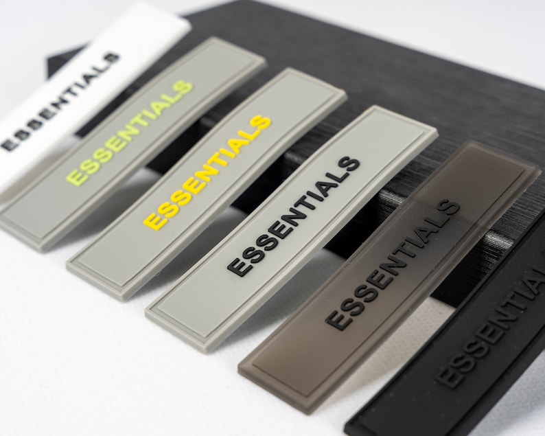 May include: Six gray fabric patches with the word "ESSENTIALS" printed in black and yellow lettering. The patches are arranged in a row, with the largest patch at the top and the smallest patch at the bottom.