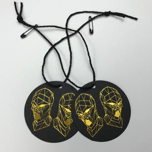 Pu&ograve; includere: Tre etichette regalo rotonde nere con disegni di viso geometrico in foglia d'oro. Ogni etichetta ha uno stringa nera attaccata con uno spillo di sicurezza.