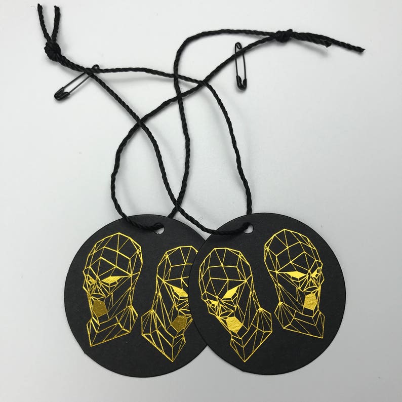 Pu&ograve; includere: Tre etichette regalo rotonde nere con disegni di viso geometrico in foglia d'oro. Ogni etichetta ha uno stringa nera attaccata con uno spillo di sicurezza.