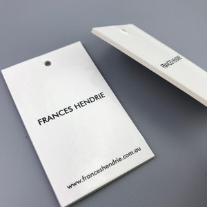 May include: Two white rectangular hang tags with rounded corners and a hole punched in the top. The tag on the left has the text "FRANCES HENDRIE" printed in black and the text "www.franceshendrie.com.au" printed in black below. The tag on the right has the text "FRANCES HENDRIE" printed in black.
