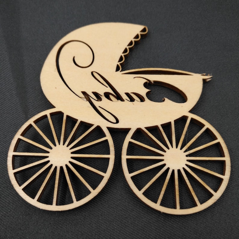 May include: Wooden cutout of a baby carriage with the word "baby" written on it. The carriage has wheels with spokes.