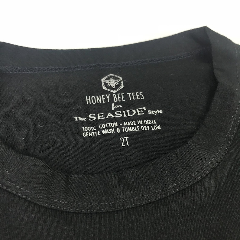 May include: Black tag with white text that reads "HONEY BEE TEES for The SEASIDE&reg; Style 100% COTTON - MADE IN INDIA GENTLE WASH & TUMBLE DRY LOW 2T". A bee with wings is in the top left corner of the tag.