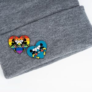 May include: A gray beanie hat with two heart-shaped pins. One pin features Mickey and Minnie Mouse embracing against a rainbow background. The other pin has the couple against a blue background. The pins are colorful and detailed.