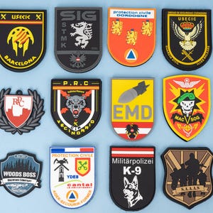 Op de afbeelding: Een verzameling geborduurde patches met diverse ontwerpen en tekst. De patches tonen emblemen, logo's en tekst zoals "USECK", "SIG", "EMD" en "WOODS BOSS BREWING COMPANY". De patches zijn in verschillende vormen en kleuren.