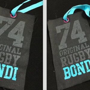 Puede incluir: Una etiqueta de tela negra con una cinta azul atada. La etiqueta tiene el n&uacute;mero "74" impreso en gris, seguido de las palabras "ORIGINAL RUGBY BONDI" en gris y azul.