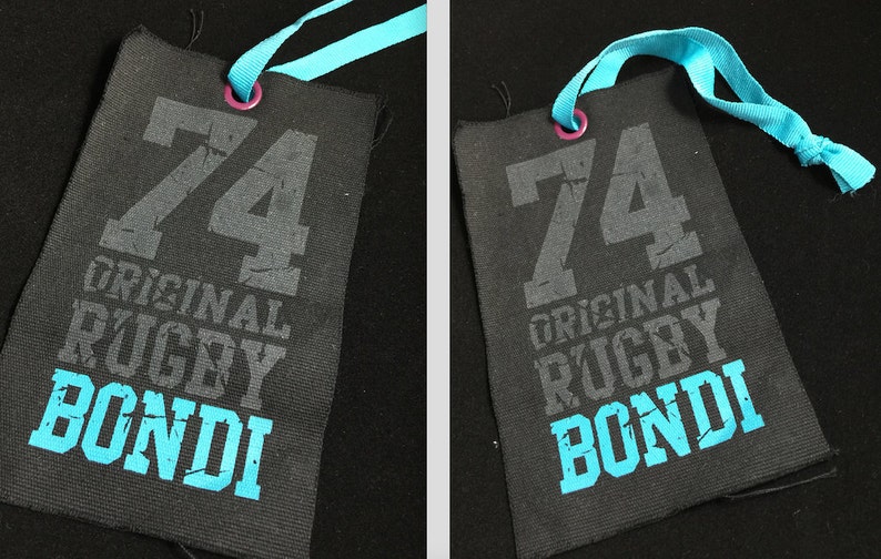 Puede incluir: Una etiqueta de tela negra con una cinta azul atada. La etiqueta tiene el n&uacute;mero "74" impreso en gris, seguido de las palabras "ORIGINAL RUGBY BONDI" en gris y azul.