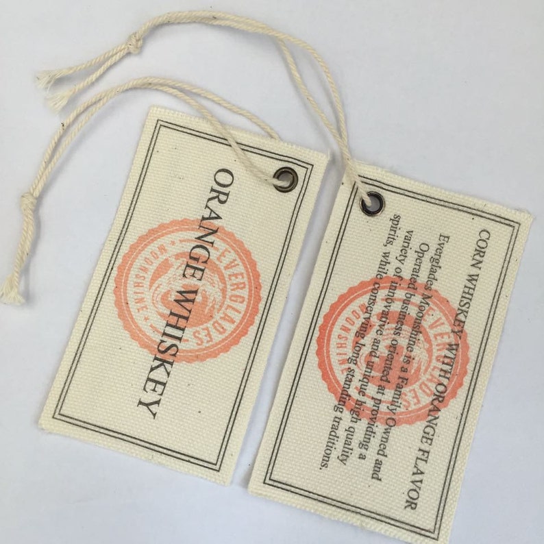 Puede incluir: Dos etiquetas de tela blanca con lazos de cuerda marr&oacute;n. La etiqueta de la izquierda dice "ORANGE WHISKEY" con un logotipo rojo y blanco. La etiqueta de la derecha dice "CORN WHISKEY WITH ORANGE FLAVOR" con un logotipo rojo y blanco.