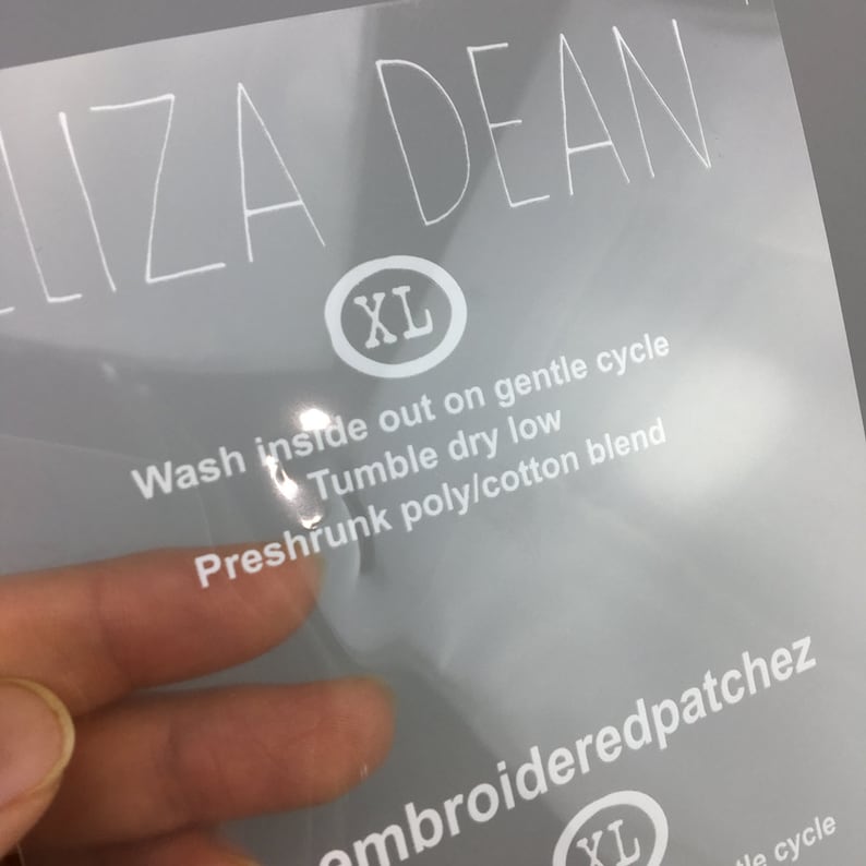 May include: White text on a clear plastic label with the words "LIZA DEAN" and "XL" in a circle. The label also includes care instructions: "Wash inside out on gentle cycle, Tumble dry low, Preshrunk poly/cotton blend".